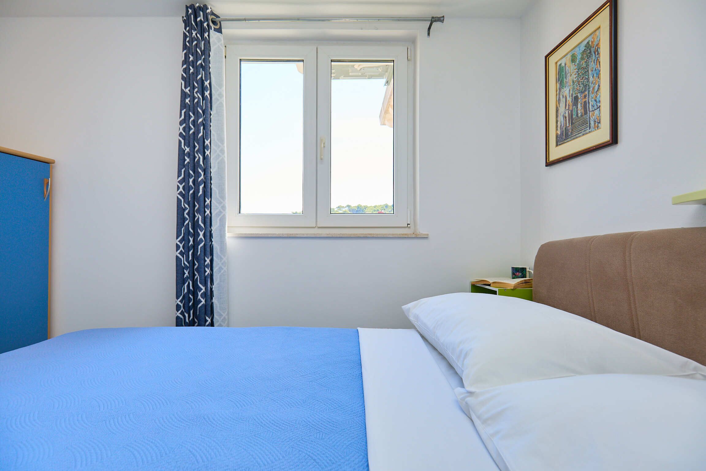 A bedroom with a bed, a window, a curtain, a framed picture on the wall, and a nightstand with a book.