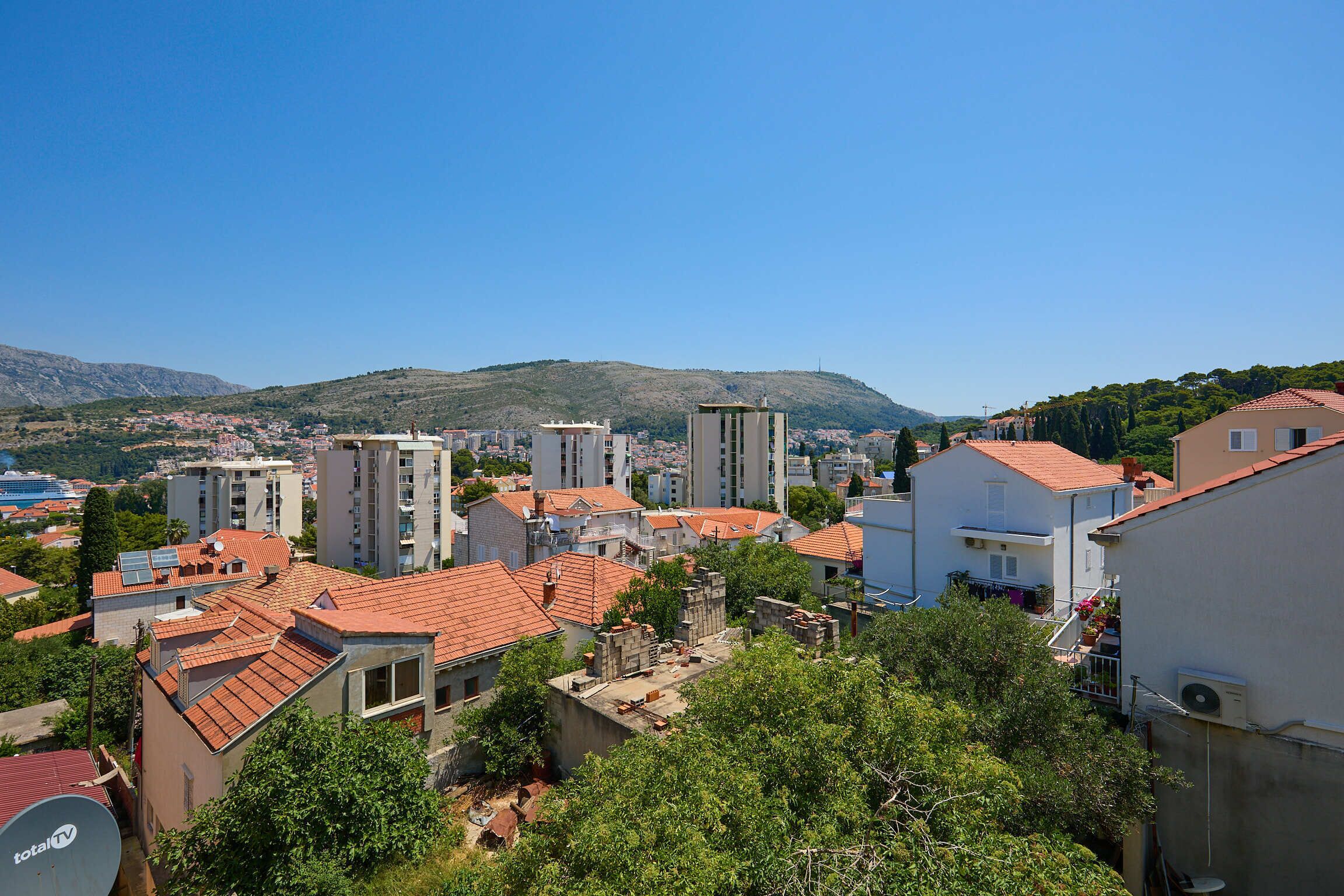 A residential area with buildings, some with red-tiled roofs, and a hill in the background.