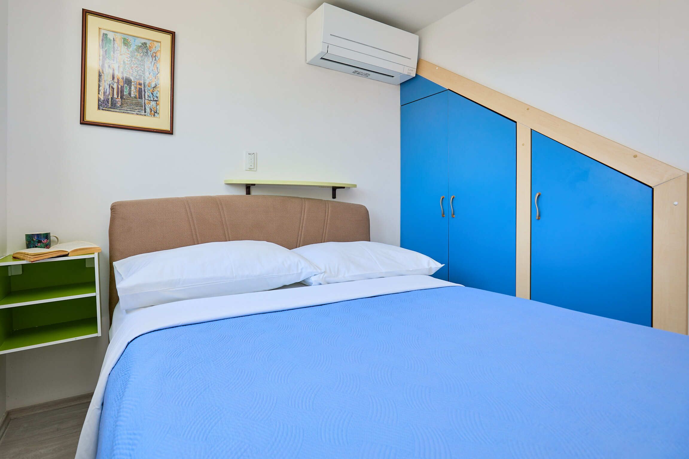 Quarto com cama, mesa de cabeceira, prateleira verde, quadro emoldurado e armários azuis.