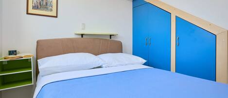 Quarto com cama, mesa de cabeceira, prateleira verde, quadro emoldurado e armários azuis.