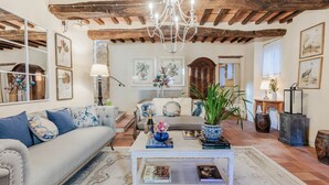 A cozy living room with a rustic ceiling, a chandelier, a sofa with blue pillows, a coffee table with books, and a large window with artwork. - Dimora delle Camelie, a traditional stylish stone farmhouse with garden on the hills of Compitese between Lucca and Pisa (Pieve di Compito)