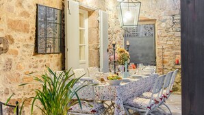 An outdoor dining area with a stone wall backdrop, a table set with plates, glasses, and a bowl of fruit, surrounded by white chairs with floral cushions. - Dimora delle Camelie, a traditional stylish stone farmhouse with garden on the hills of Compitese between Lucca and Pisa (Pieve di Compito)