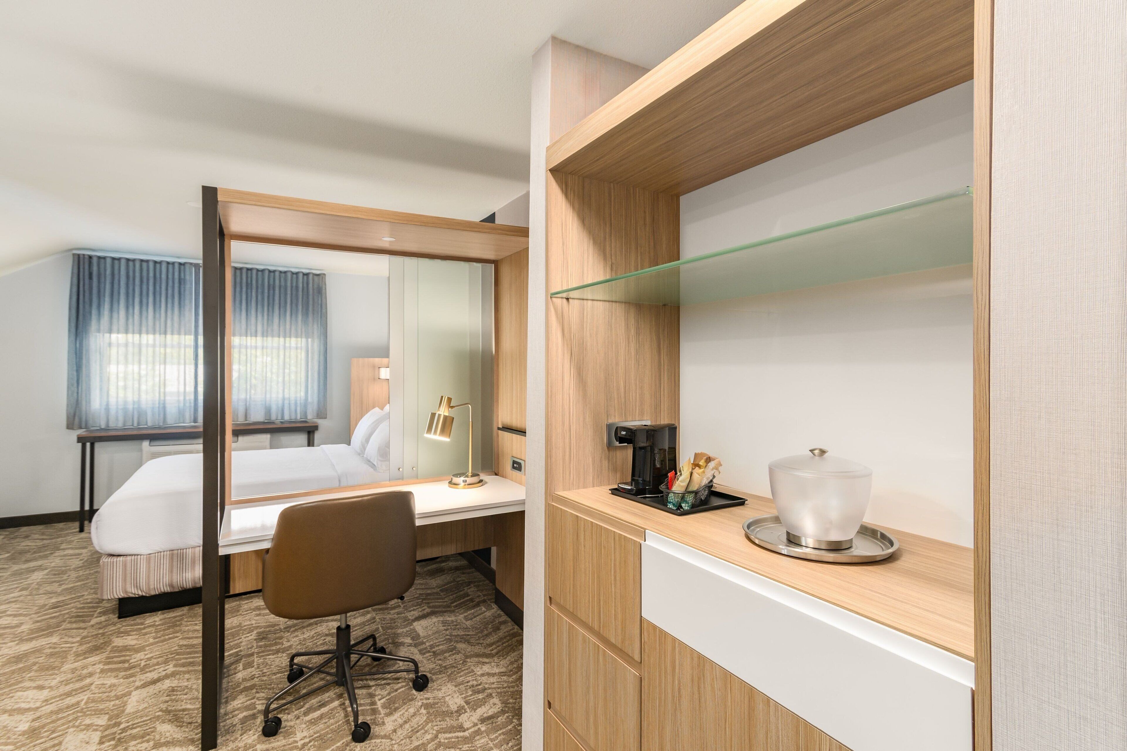 A hotel room with a bed, desk, chair, and a coffee machine.