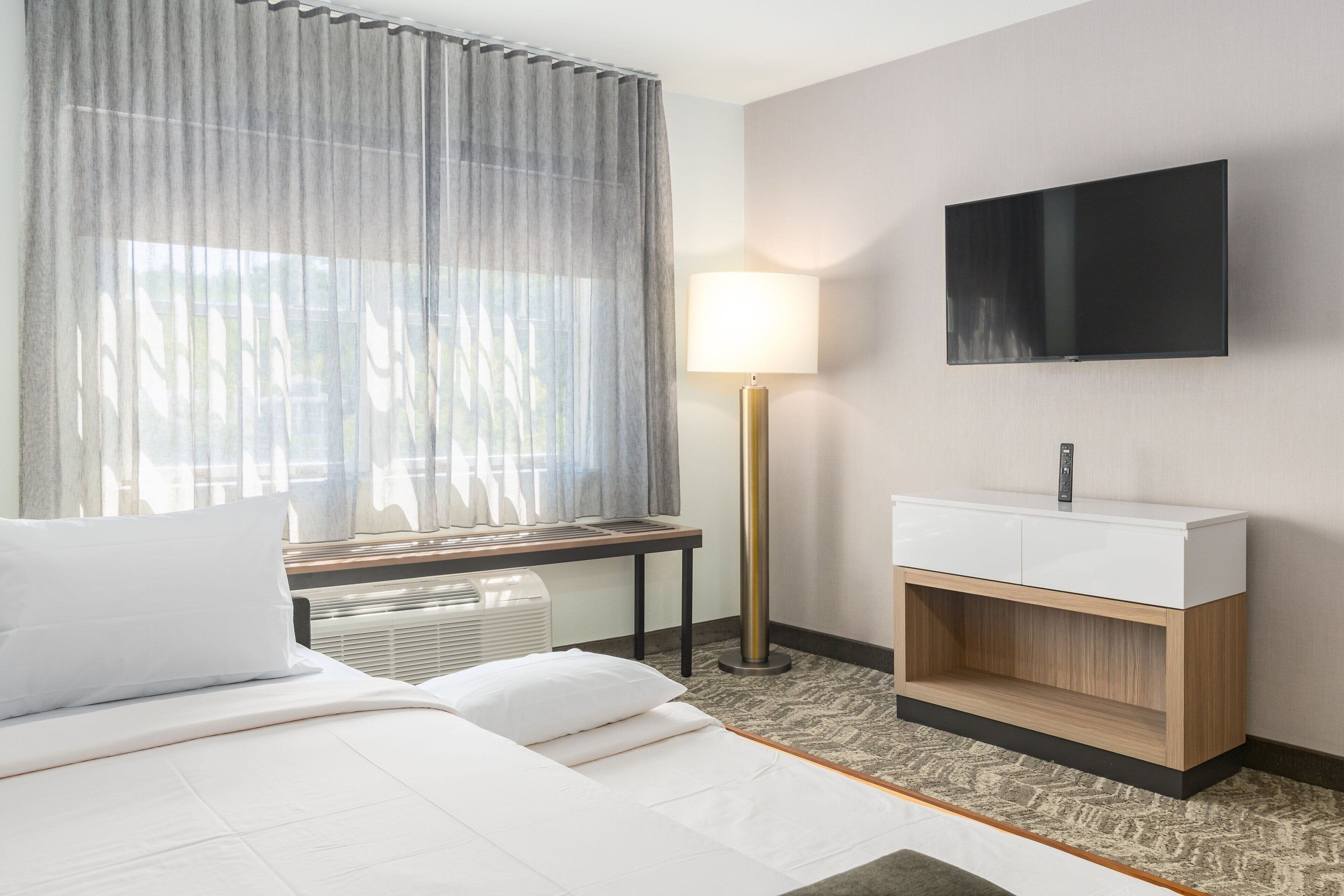 A hotel room with a bed, a bedside table, a lamp, a TV mounted on the wall, and a window with sheer curtains.