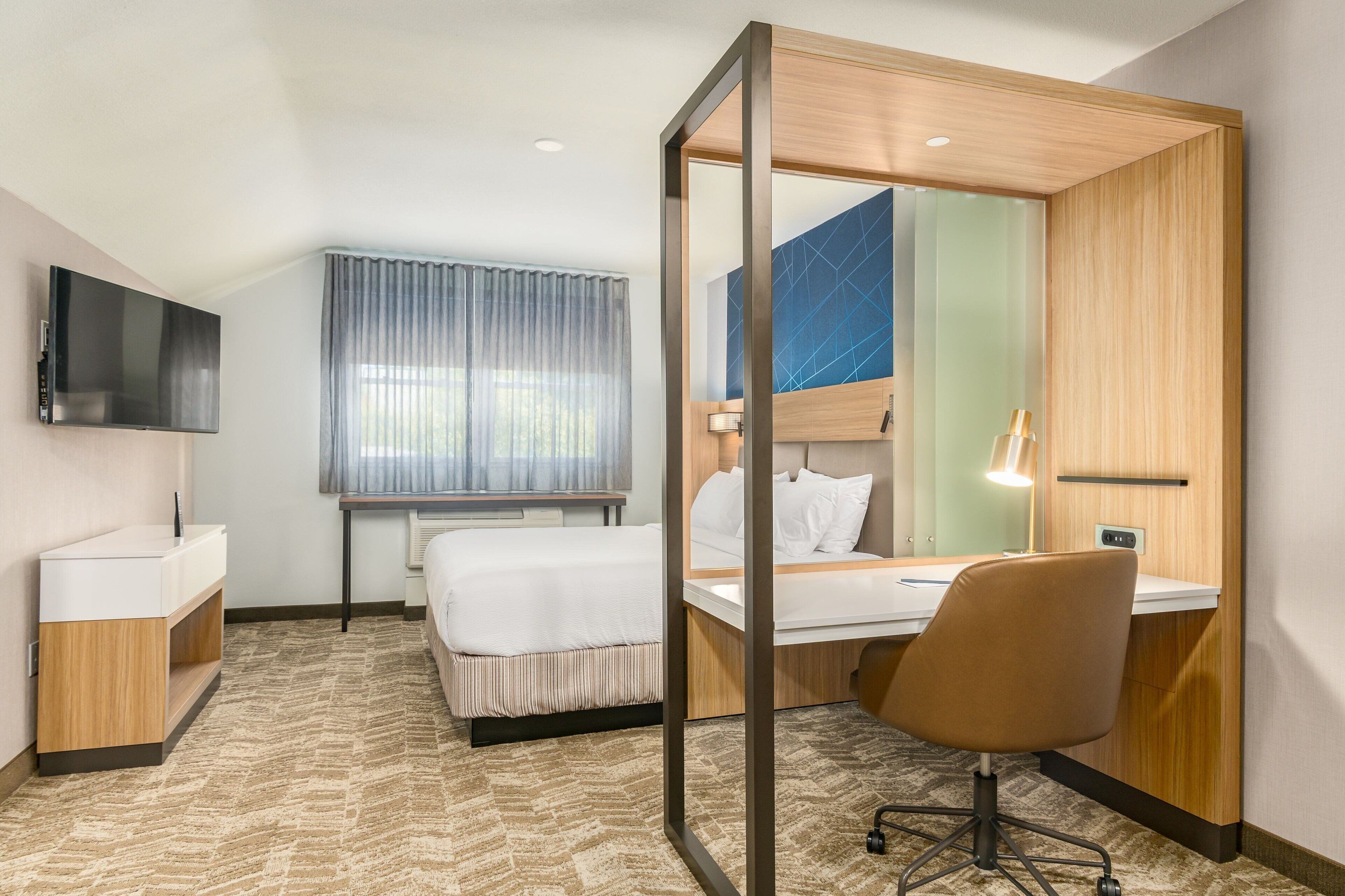 Ein Hotelzimmer mit Bett, Schreibtisch, Stuhl, Fernseher, Spiegel und einem Fenster mit Vorhängen.