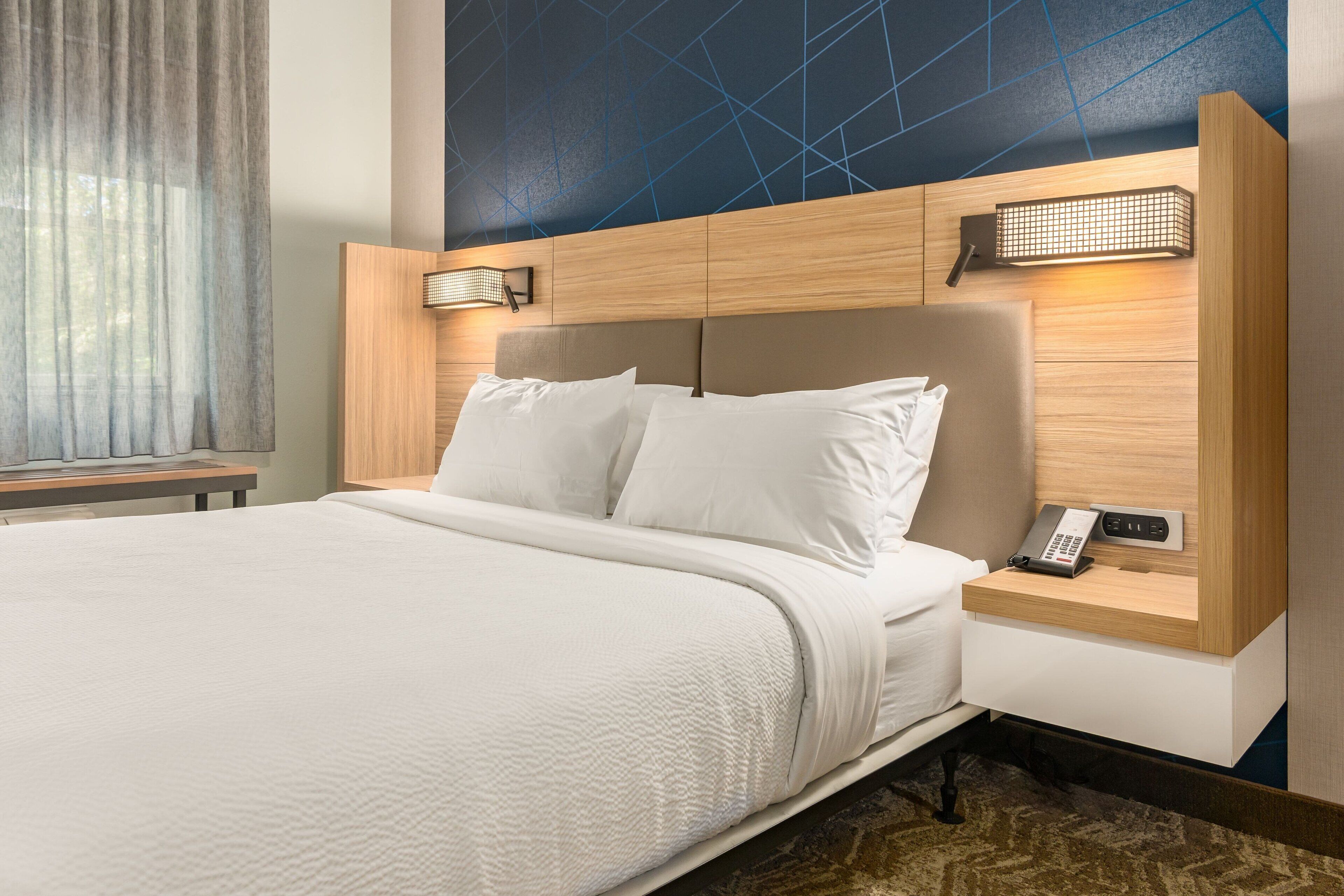 Een net opgemaakt bed met witte lakens, een houten hoofdbord en een nachtkastje met een telefoon en een lamp.