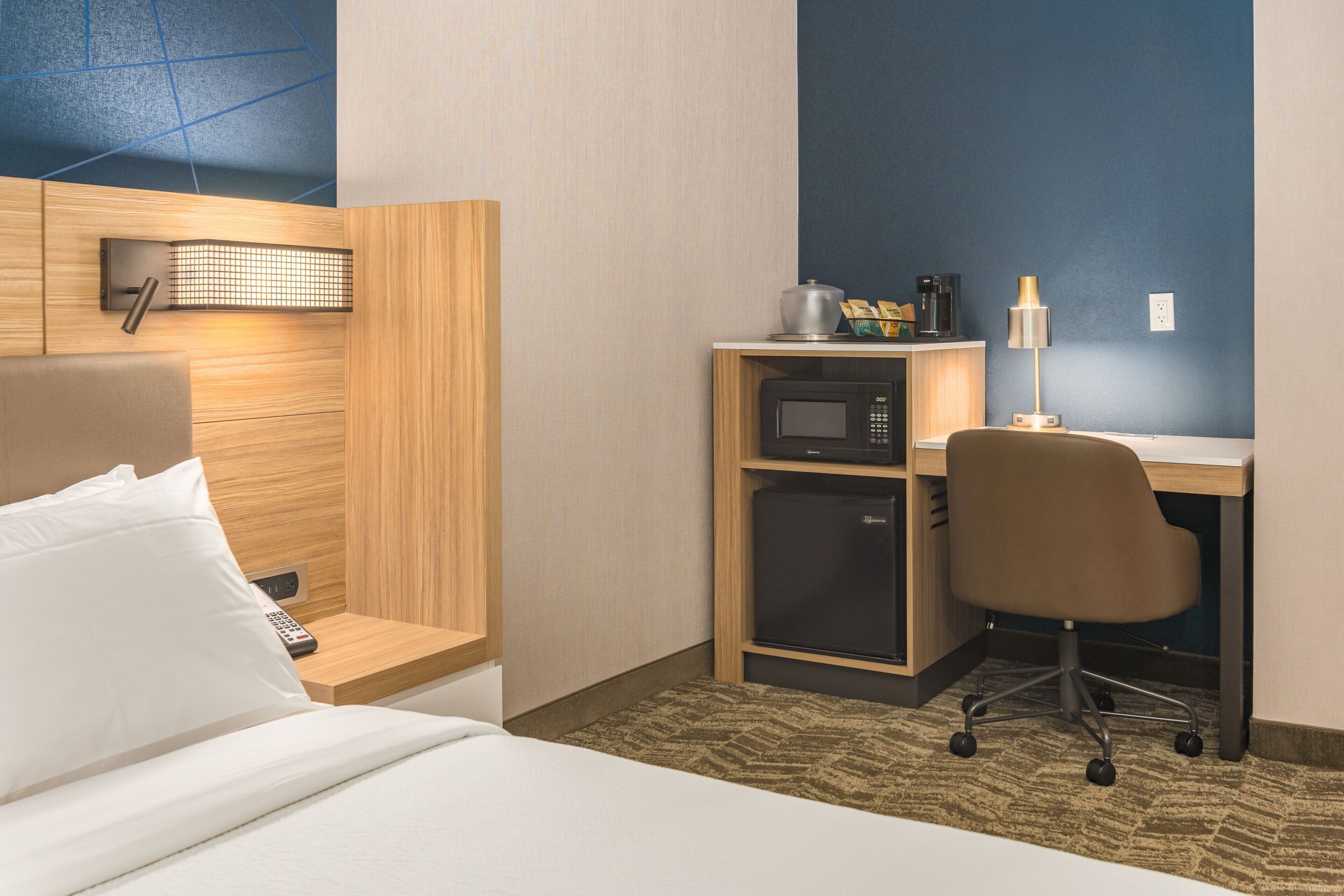 A hotel room with a bed, a desk with a computer, a microwave, a coffee maker, and a lamp.