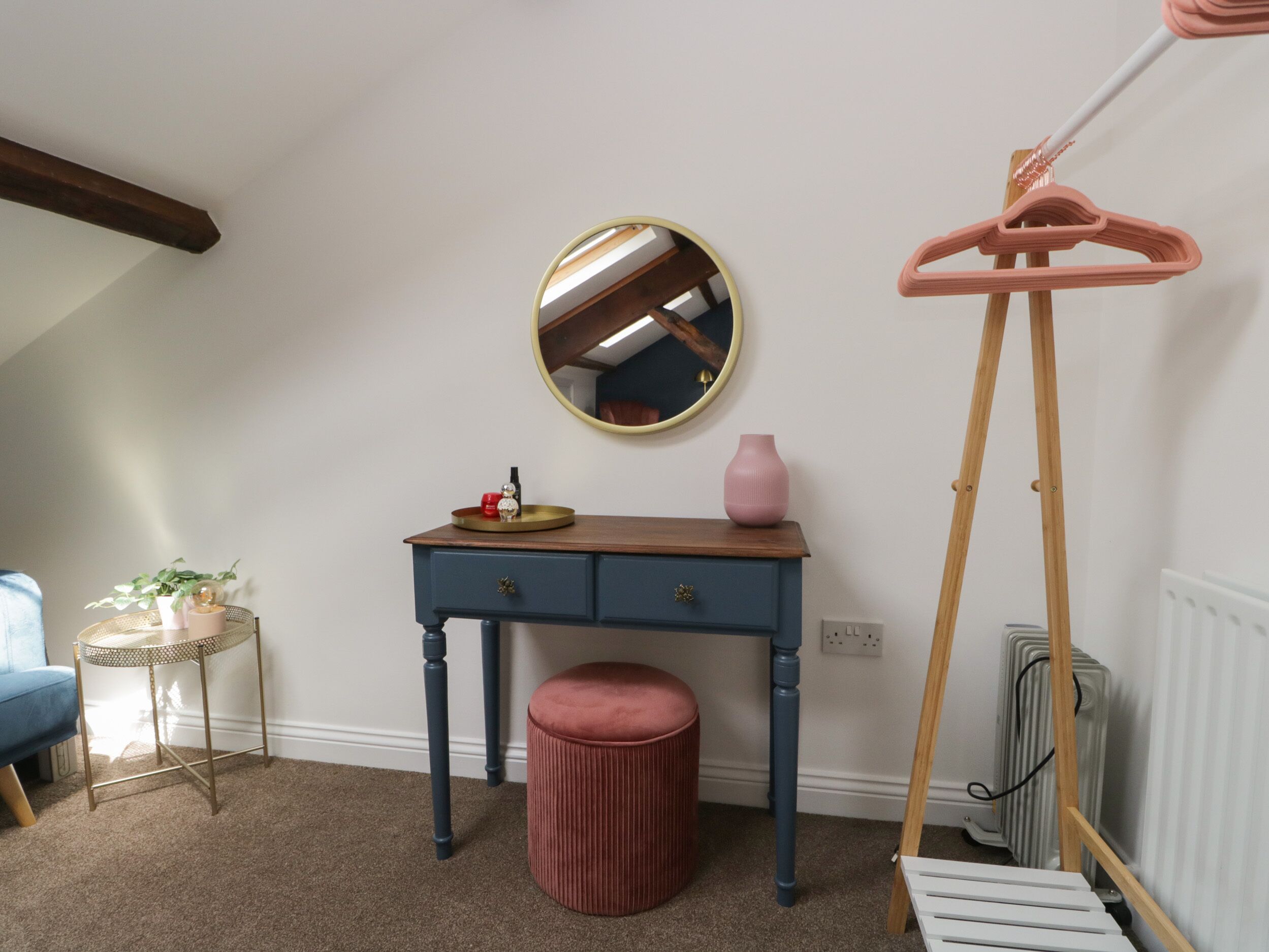 Een slaapkamer met een houten dressoir, een spiegel, een roze vaas, een rode kruk, een houten kapstok en een radiator.