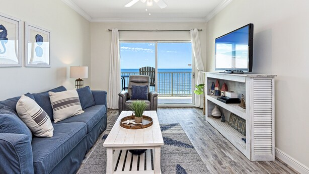 A living room with a sofa, coffee table, TV, and a balcony with a view of the ocean. - Crystal Shores 606 | Unforgettable Gulf Front Views | Indoor & Outdoor Pools (Gulf Shores)