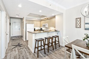 A modern kitchen with wooden cabinets, a central island with stools, and a dining table with chairs. - Crystal Shores 606 | Unforgettable Gulf Front Views | Indoor & Outdoor Pools (Gulf Shores)