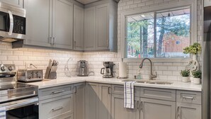 Ett modernt kök med gråa skåp, rostfria vitvaror och ett fönster med utsikt över träd och en byggnad.
