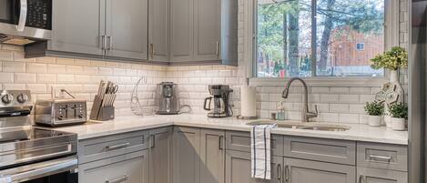 A modern kitchen with grey cabinets, stainless steel appliances, and a window with a view of trees and a building.