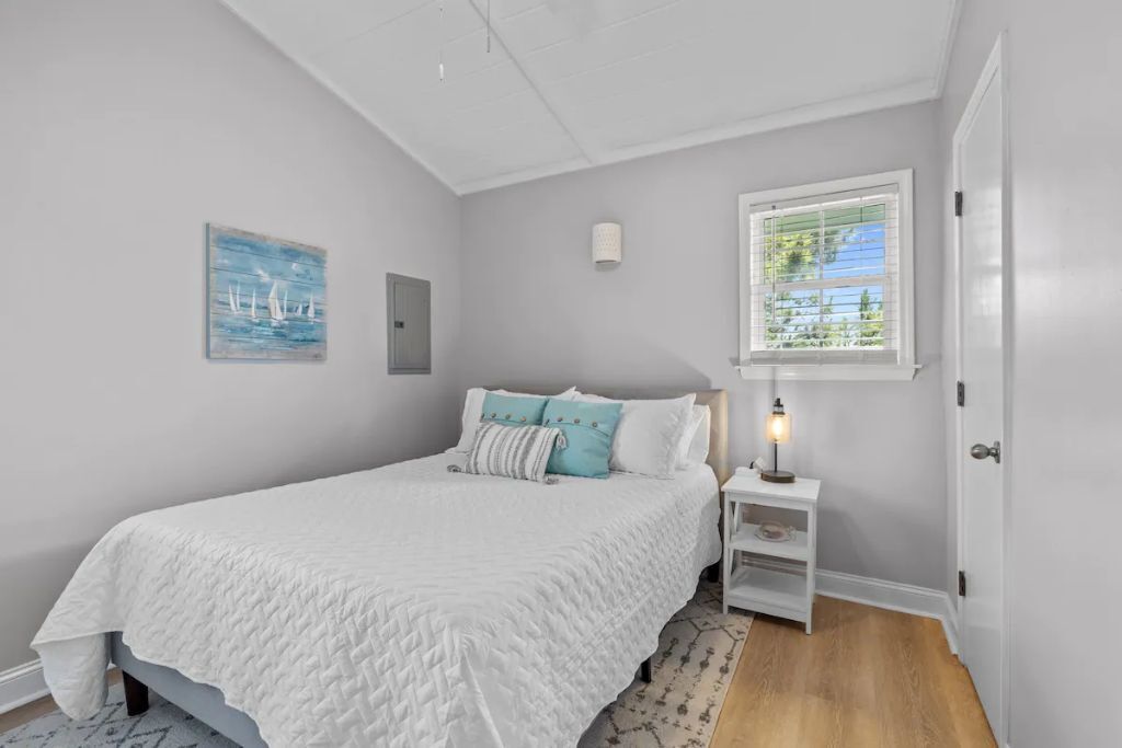 A bedroom with a bed, a nightstand, a lamp, a window with blinds, and a painting on the wall.