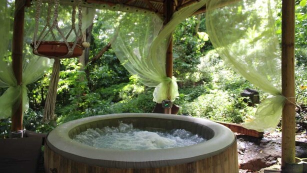 A hot tub in a covered outdoor area with hanging macramé decorations and a lush green background. - ecolodge Martinique (Fort-de-France)