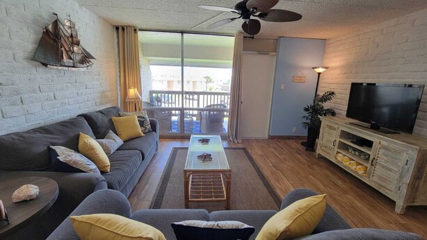 A living room with a sofa, coffee table, TV, and a balcony with a view. - King 1bd/1ba Condo w/ Pool View & Beach Access (Port Aransas)