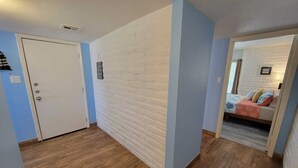 A room with a white brick wall, a wooden floor, a bed with a blue and orange bedspread, and a door with a small shelf above it. - King 1bd/1ba Condo w/ Pool View & Beach Access (Port Aransas)