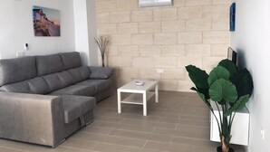 Una sala de estar con un sofá gris, una mesa de centro blanca, una planta verde grande y un ventilador de techo.