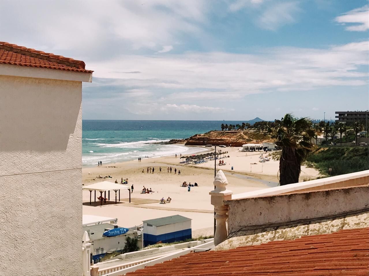 Vista da praia a partir de uma varanda com uma parede branca, uma palmeira e um afloramento rochoso ao longe.