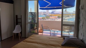 A hotel room with a bed, a chair, a small table, and a balcony with a view of the sea and buildings. - RIOPASSIONS, LARGE T3, BEAUTIFUL SEA VIEW (Toulon)