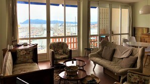 A spacious living room with a large window offering a view of a marina and cityscape. There are two sofas, a coffee table, and a dining table with chairs. - RIOPASSIONS, LARGE T3, BEAUTIFUL SEA VIEW (Toulon)