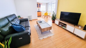 Un salon moderne avec un canapé en cuir noir, une table basse, un téléviseur à écran plat et un coin repas avec des chaises jaunes.