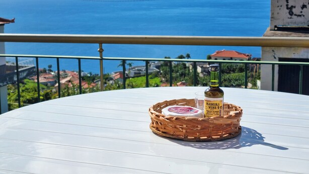 Une table ronde blanche sur laquelle se trouve un panier en osier contenant une bouteille de vin de Madère et un verre. La table est placée sur un balcon d’où l’on peut admirer la mer et les bâtiments qui se dressent à l’arrière-plan.