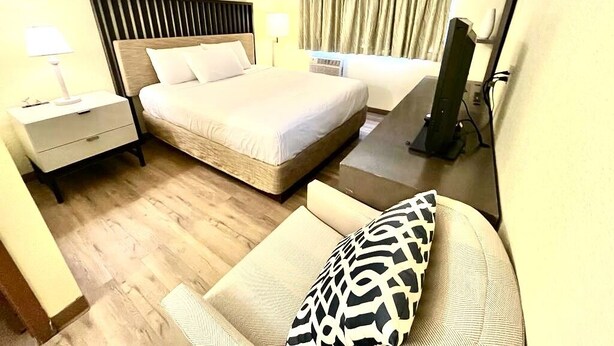 Ein Hotelzimmer mit einem großen Bett, einem Nachttisch mit Lampe, einem Sessel mit gemustertem Kissen und einem Fernseher.