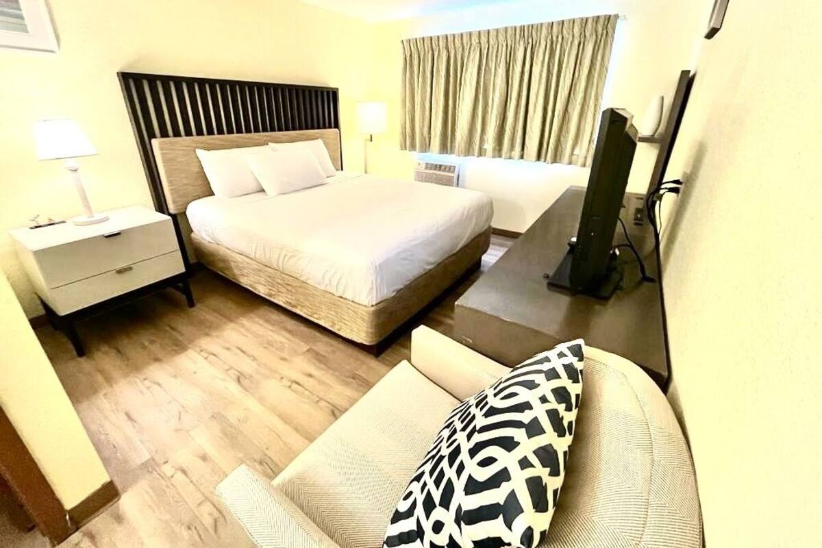 A hotel room with a large bed, a bedside table with a lamp, a chair with a patterned cushion, and a television.