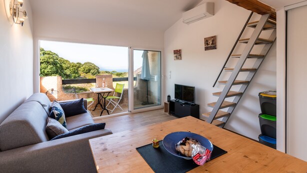 A modern living room with a wooden table, a sofa, a TV, and a sliding glass door leading to an outdoor area. - Ambroggio - Grand studio avec mezzanine - Vue Mer (Lumio)