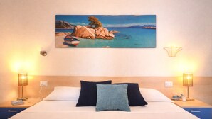 A hotel room with a bed, two bedside lamps, a painting of a coastal scene, and a nightstand with a lamp and a cup. - Libeccio | Relax Terrace 30m from the beach (Isola Rossa)