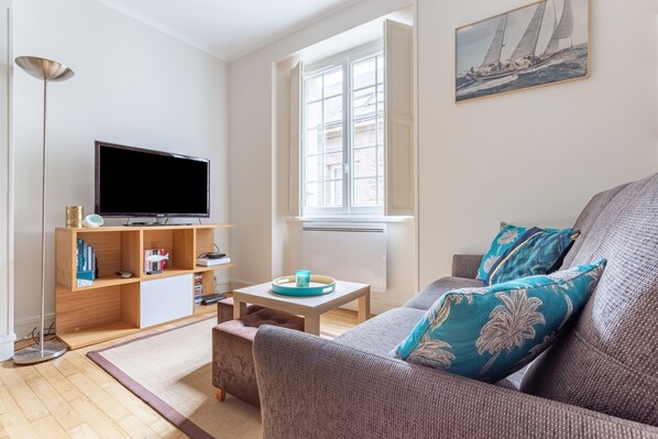 A living room with a flat-screen TV, wooden shelving, a small table with a tray, a sofa with patterned cushions, and a window with a sailboat painting.