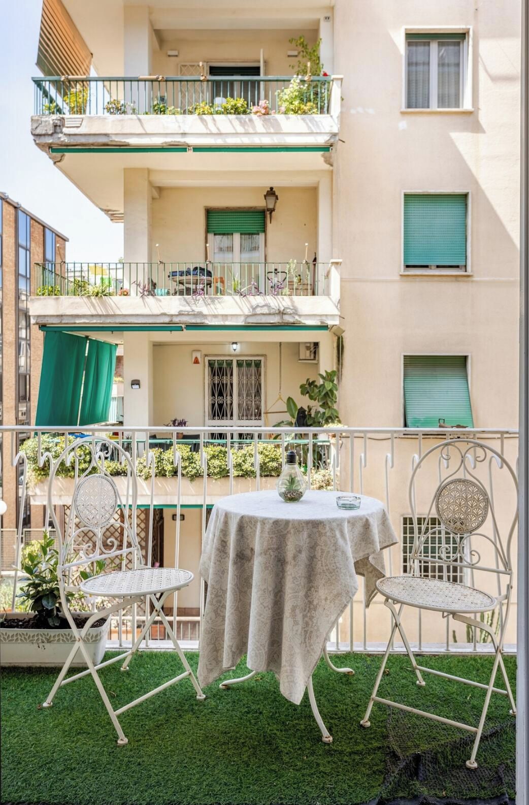 A balcony with a table and chairs, surrounded by greenery and a building facade.