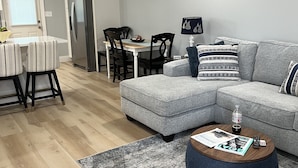 Un salon moderne avec un canapé d’angle gris, une table basse ronde et un coin repas comprenant une table blanche et des chaises à rayures.