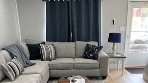 Un salon avec un canapé d’angle gris, une table basse ronde en bois et un rideau bleu foncé.