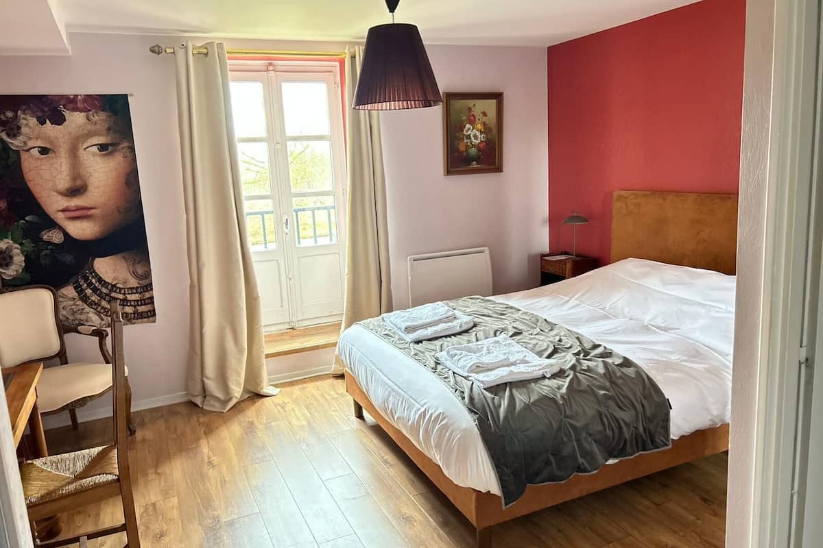 A bedroom with a bed, a chair, a desk, a painting, a window, and a radiator.