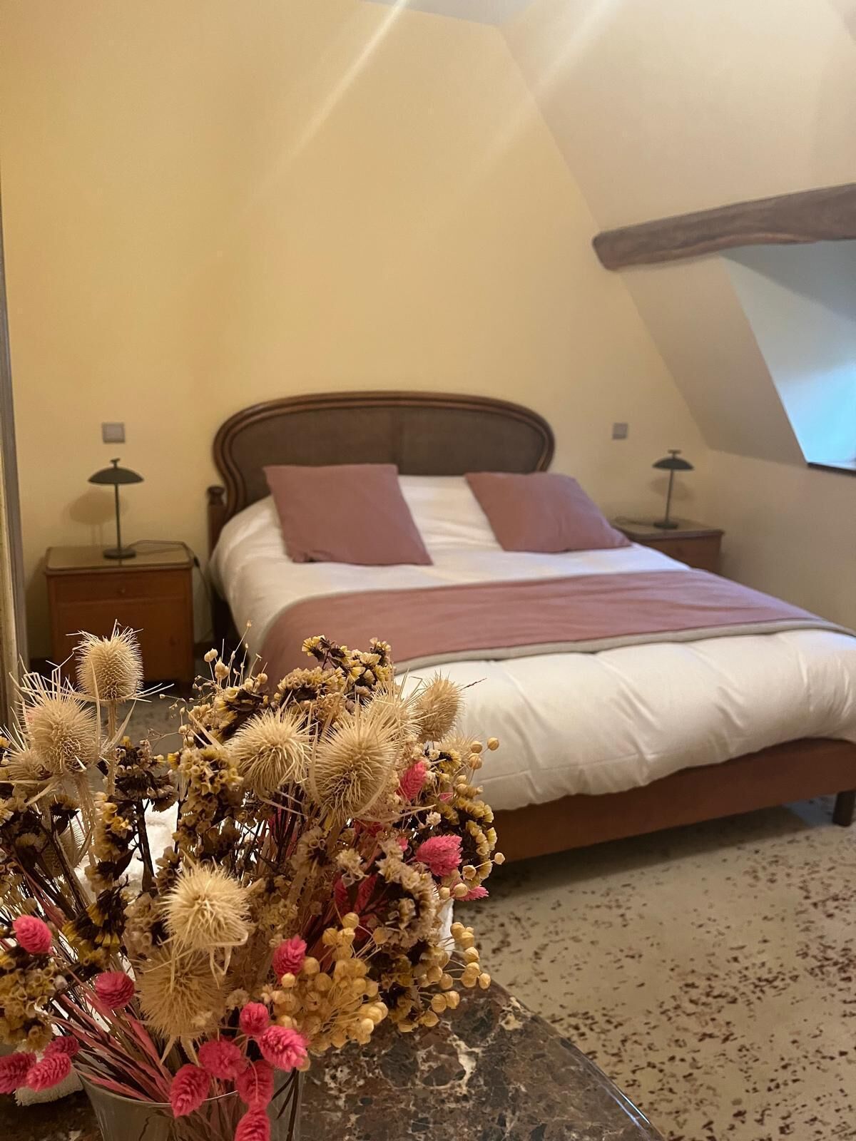 Une chambre à coucher avec un lit, des oreillers, une table de chevet, une lampe et un vase contenant des fleurs séchées.