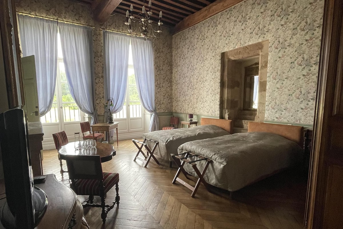 A room with a wooden herringbone floor, two beds with headboards, a dining table with chairs, a chandelier, and a large mirror on the wall.