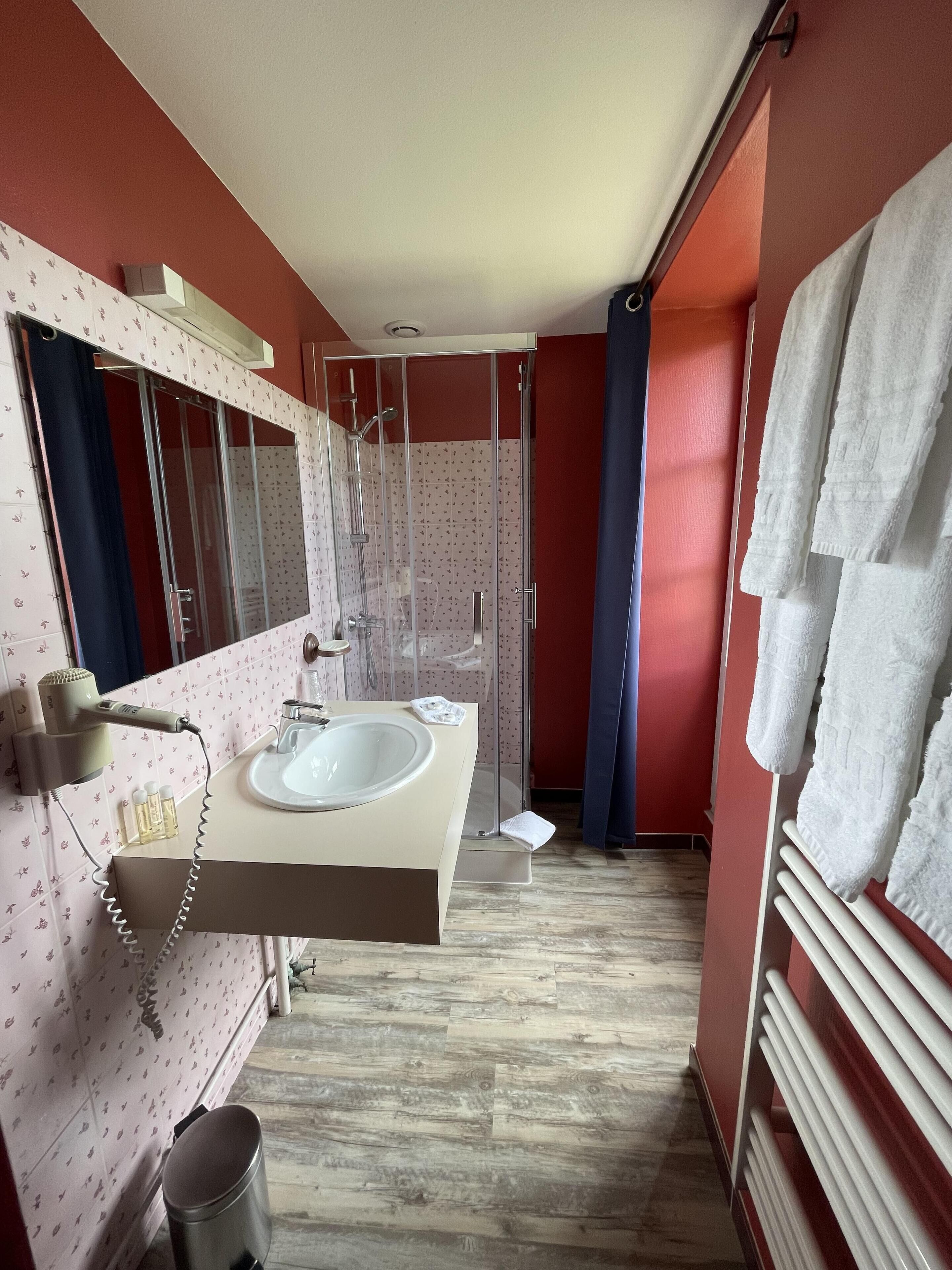 A bathroom with a large mirror, a sink, and a shower. There are towels hanging on a rack and a trash can.