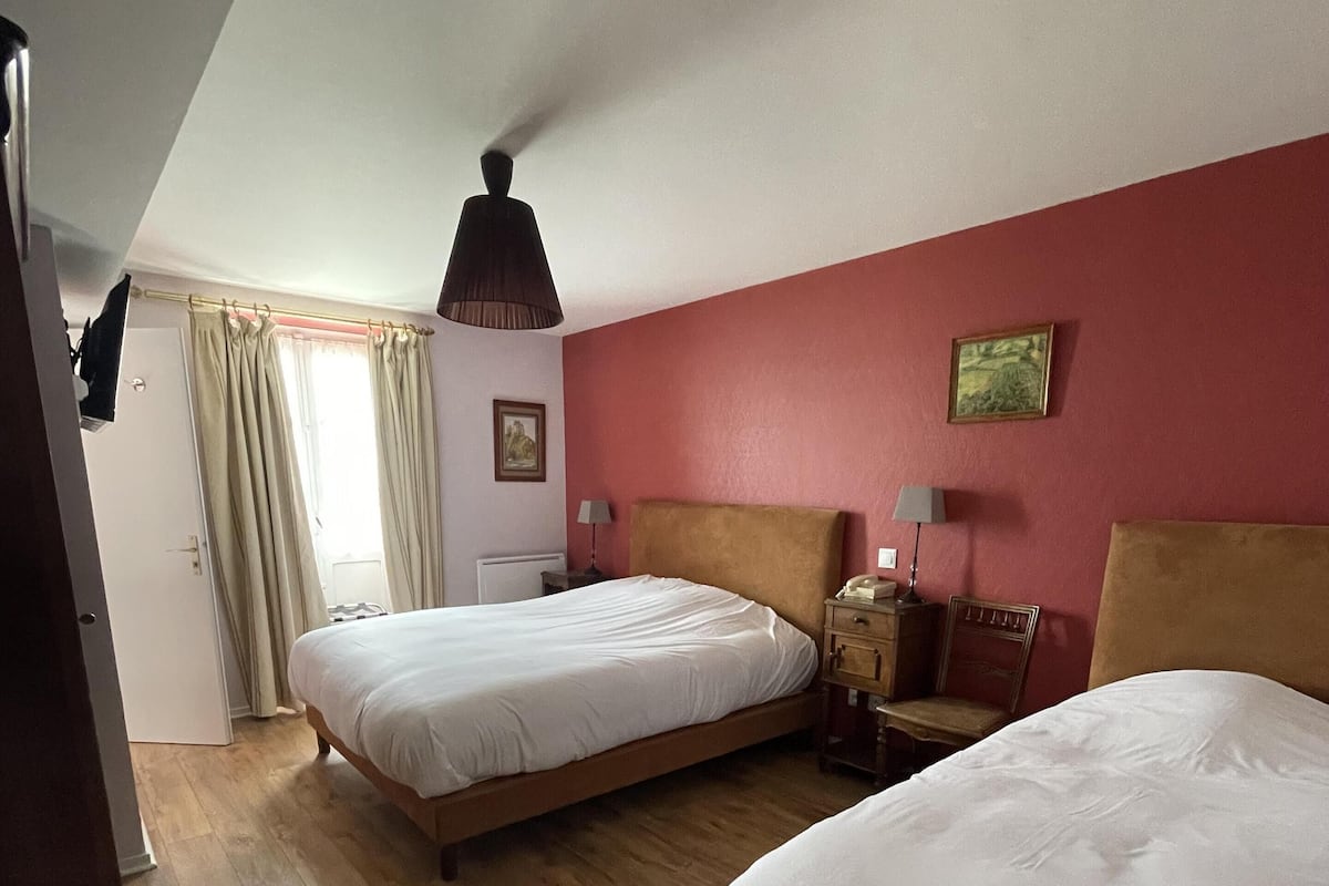 A hotel room with two beds, wooden flooring, a red accent wall, a ceiling lamp, and a window with curtains.