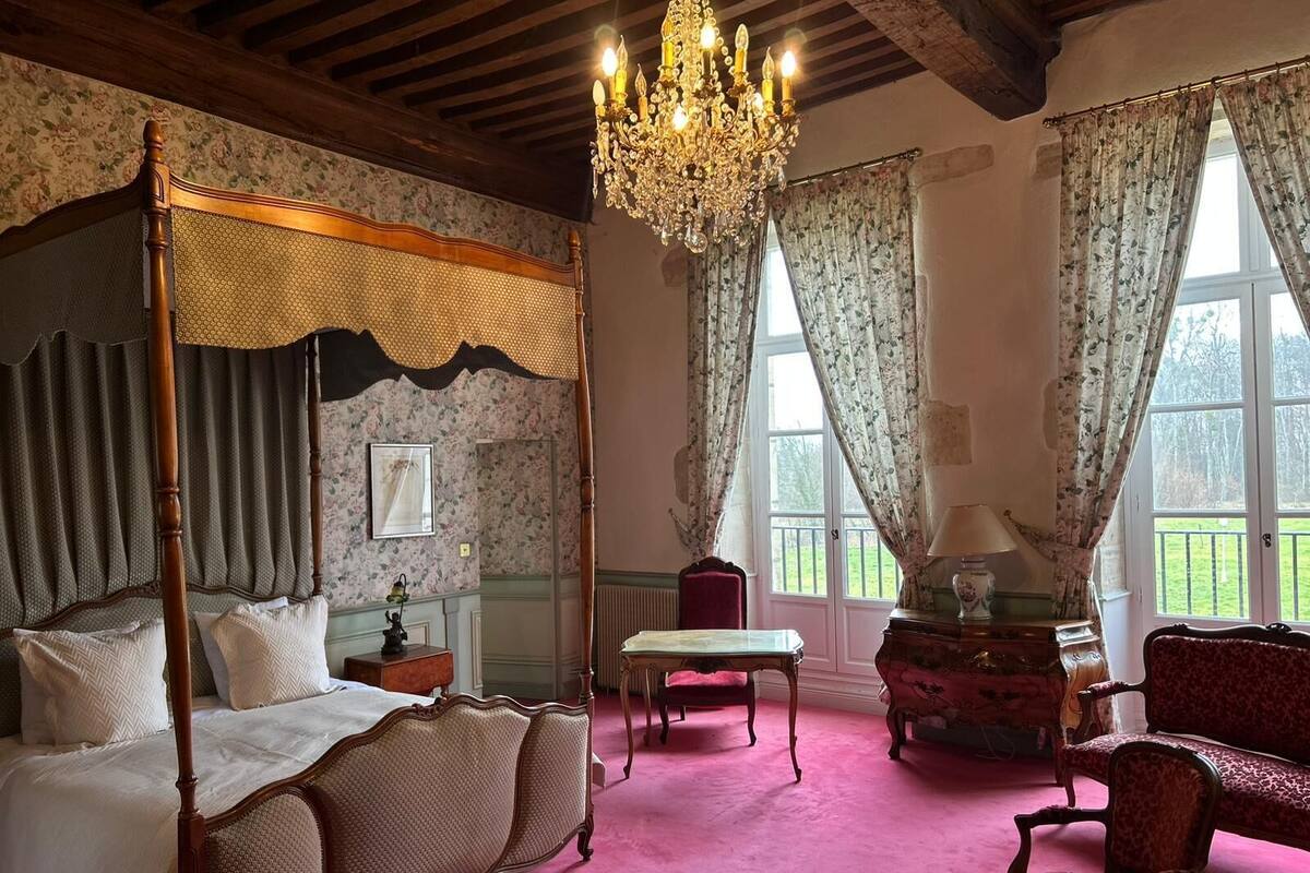 A four-poster bed with a canopy, a chandelier, a red armchair, a wooden dresser, and a view of the garden through large windows.