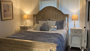 A bedroom with a large bed, two bedside tables, a nightstand, and a window with blinds. - Tyler's Texas Rose, a beautiful 2BR/1BA house with back yard (Tyler)