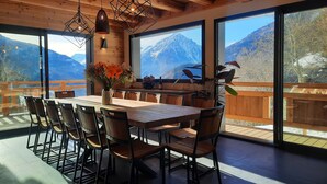 Un espace repas avec une table et des chaises en bois, de grandes fenêtres offrant une vue sur les montagnes et des lampes suspendues.