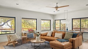 A spacious living room with a brown leather sofa, a round coffee table, and a dining area with a table and chairs. Large windows provide a view of the outdoors.