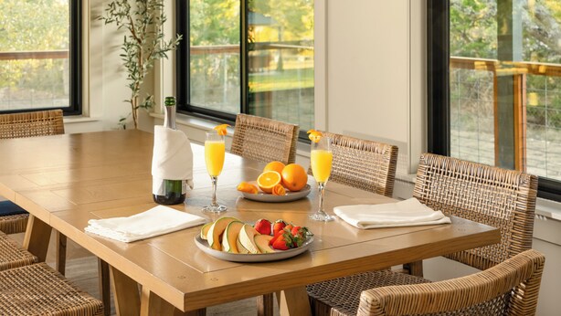A dining area with a wooden table, wicker chairs, a bottle of wine, glasses of orange juice, and a plate of fruit.