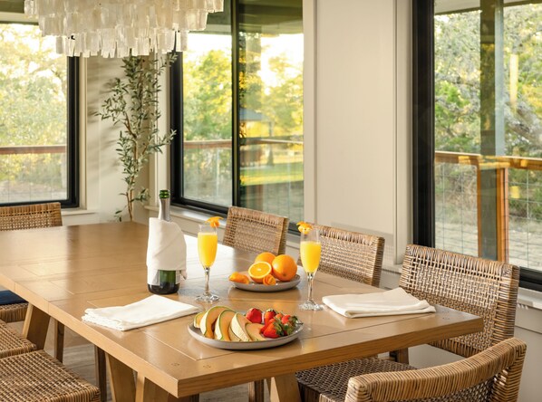 A dining area with a wooden table, wicker chairs, a bottle of wine, glasses of orange juice, and a plate of fruit.