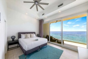 A bedroom with a bed, a nightstand, a ceiling fan, and a large window with a view of the ocean.