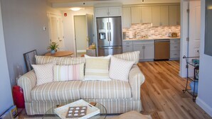 A living room with a striped sofa, a glass coffee table, and a kitchen with wooden flooring and white cabinets. - Brand new Ocean View Condo with private balcony, steps from the beach! (Dennis Port)