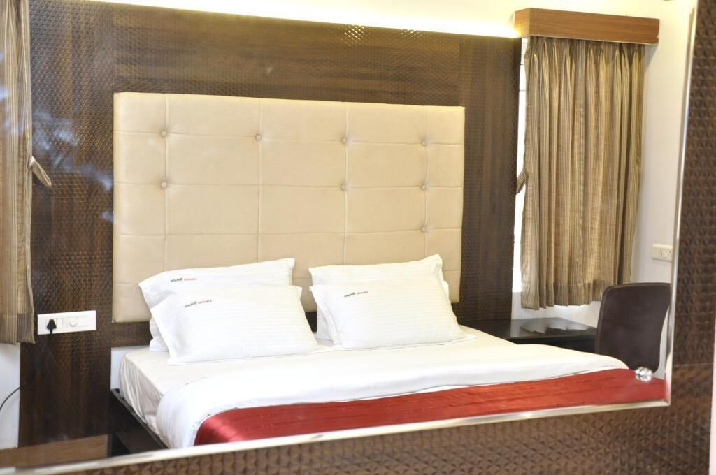 Une chambre d’hôtel dotée d’une grande tête de lit capitonnée, d’un lit avec du linge de lit blanc, d’un élément décoratif rouge au pied du lit et d’une vue sur l’extérieur par une fenêtre ornée de rideaux.