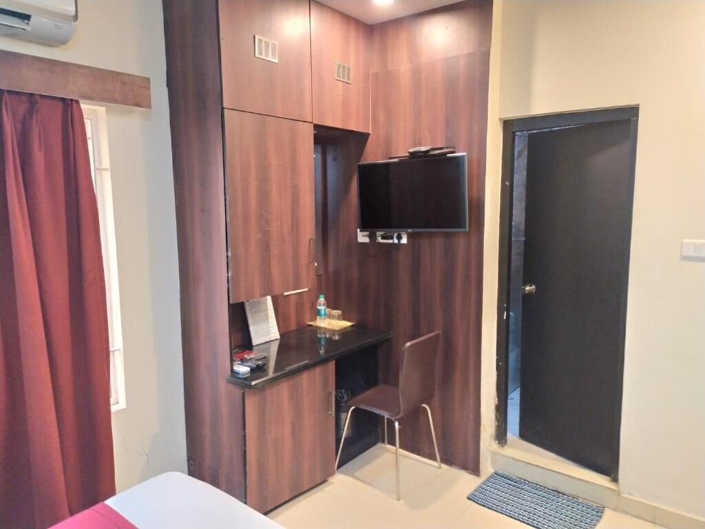 A compact room with a bed, a desk with a chair, a TV mounted on the wall, and a door.