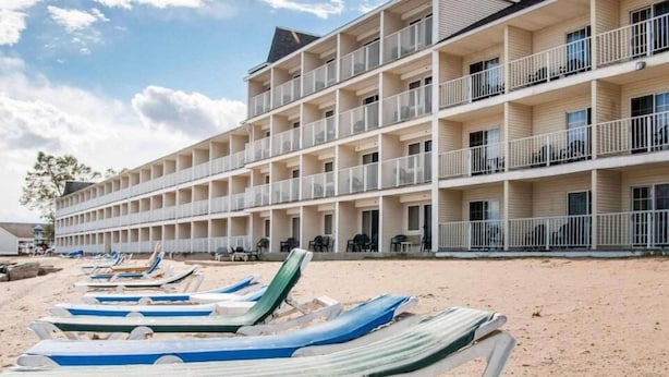 A multi-story hotel with balconies, a sandy beach in front, and lounge chairs. - Canberra Premiere Comfort Hotel (Mackinaw City)